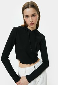 Sudadera recortada negra con textura acanalada, dobladillo con forma de onda y cordón. Las mangas largas terminan en puños con volantes. Se lleva con jeans blancos de cintura alta.