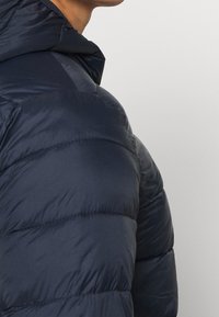 Zimní bunda v tmavě námořnické modři, vyrobená z lesklé prošívané látky. Obsahuje kapuci a šité panely pro izolaci a teplo.