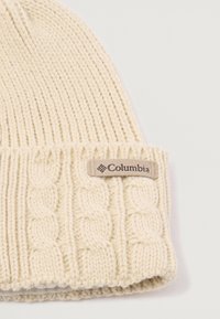 Kootud müts kreemjas valges värvuses, millel on sooniktekstuur ja kabli kudumise muster. Küljel on pruun Columbia brändi silt.