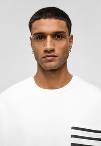 Witte sweatshirt met een ronde hals en korte mouwen. Heeft een zwarte gestreepte zak aan de linkerkant. Gladde textuur, casual ontwerp.