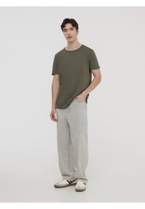 T-shirt verde oliva a maniche corte abbinato a jeans larghi grigi chiari e sneakers bianche con accenti marroni. Design semplice e casual.