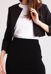 Veste noire courte portée sur un t-shirt blanc à col rond, associée à une jupe noire ajustée texturée. Design sur mesure avec une structure minimaliste.