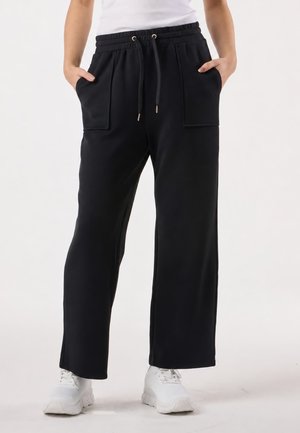 Personne portant un pantalon noir à jambes larges avec poches plaquées à l'avant et taille à cordon, debout, les mains dans les poches, et des baskets blanches.