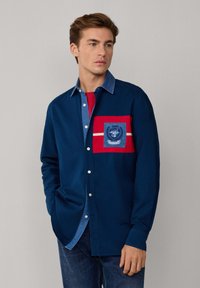 Camicia a maniche lunghe blu navy con tasca patch rossa, caratterizzata da dettagli in denim blu e un logo. Chiusura frontale con bottoni e colletto a contrasto.