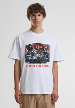 Jeune homme portant un t-shirt blanc Nirvana "Live in New York" avec un motif coloré d'une performance scénique, debout devant un fond uni.