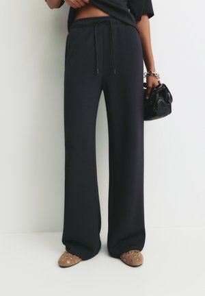 Person trägt lockere schwarze Hosen, hält eine kleine schwarze Handtasche und trägt braune flache Schuhe mit silbernen Ösendetails.