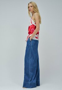 Mujer con cabello largo y rubio, vistiendo una camiseta blanca de tirantes con estampado floral rojo y unos vaqueros de tela vaquera azul de pierna ancha, de pie de lado con las manos en los bolsillos.