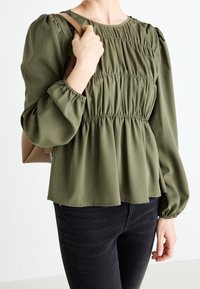 Blouse vert olive avec texture froncée, manches longues bouffantes et encolure ronde, associée à un jean noir et un sac marron clair.