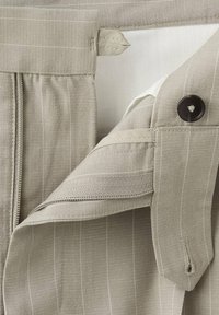 Gros plan d’un pantalon beige à fines rayures verticales, montrant les détails de la fermeture éclair, du bouton et de la taille sur un fond blanc.