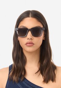 Occhiali da sole in acetato nero con montatura quadrata e lenti sfumate. Presentano astine spesse e una finitura lucida. Accompagnati da capelli castani e un outfit casual.