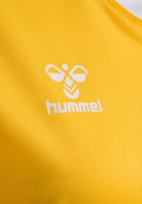 Gult atletiskt linne med slät textur, med en vit logotyp som föreställer ett bi och varumärkesnamnet "hummel" nedanför.