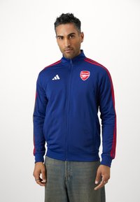 Mėlyna zip-up striukė su raudonomis juostelėmis, turinti Arsenalo logotipą ant krūtinės ir baltą Adidas logotipą ant peties. Lygi audinio tekstūra.