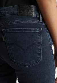 Mörkblå denimjeans med rak passform. Har en svart läderlapp på midjebandet och ett distinkt sömnadsmönster på bakfickorna.