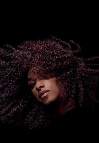 Frau mit voluminösem, lockigem, dunkelburgunderrotem Haar vor schwarzem Hintergrund, der Kopf leicht geneigt mit neutralem Gesichtsausdruck.