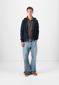 Hanorac cu fermoar de marină peste un tricou polo maro, asortat cu blugi deschiși lejeri și pantofi slip-on maro. Design simplu și casual.