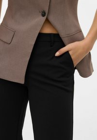 Blazer marrone con un solo bottone, abbinato a pantaloni neri su misura. Il blazer presenta un colletto a lancia; i pantaloni hanno una vestibilità slanciata con tasche.