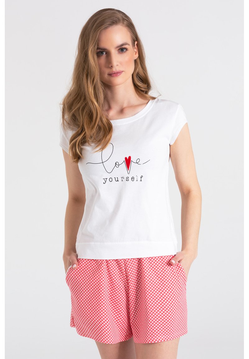 Biała bawełniana koszulka z czarnym napisem "ukończ siebie" i czerwonym sercem, w zestawie z różowymi w kratkę szortami.