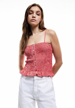 Jeune femme aux longs cheveux bruns portant un haut sans manches à carreaux rouge et blanc et un pantalon blanc sur fond blanc.