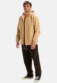 Beige waterdichte jas met capuchon, drukknoopsluiting, trekkoorden en logo. Draagover een bruine blouse en donkerbruine corduroybroek. Sneakers eronder.