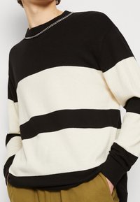 Suéter a rayas negras y crema con cuello redondo, tejido de punto, puños acanalados y detalle de cadena plateada alrededor del cuello.