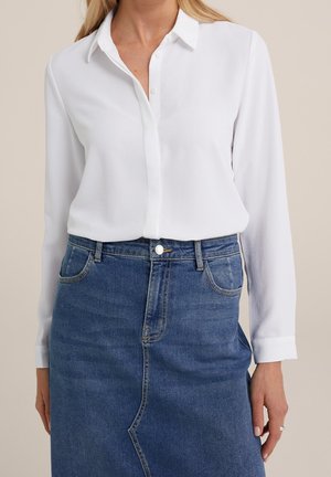 Femme portant une chemise blanche à manches longues boutonnée, rentrée dans une jupe en jean bleu taille haute avec poches avant.