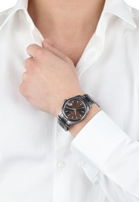 Zilveren roestvrijstalen horloge met een bruine wijzerplaat, zilveren uurmarkers en een datumfunctie, gedragen om een pols in de buurt van een witte shirtkraag.