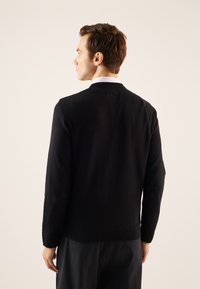 Pull noir à col rond et à manches longues. Fabriqué en tissu lisse, avec des poignets et un ourlet côtelés. Porté par-dessus un pantalon sombre.