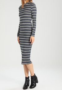 Robe à manches longues rayée bleu marine et blanche, design ajusté, texture côtelée et ourlet longueur genou. Portée avec des bottines noires.