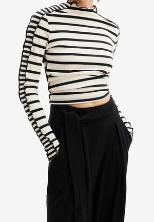 Personne portant un haut à manches longues à rayures horizontales blanches et noires et un pantalon noir taille haute avec une ceinture nouée et les mains dans les poches.