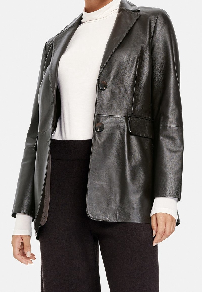 Czarna skórzana marynarka o dopasowanym kroju, z klapami z wycięciem w szpic, dwoma guzikami i bocznymi kieszeniami, noszona na białym golfie. Gładka faktura.