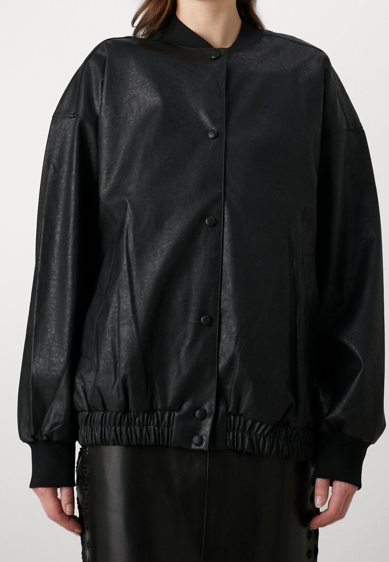 Femme portant une veste bomber en similicuir noir avec des boutons-pression et un ourlet et des poignets élastiqués, debout devant un fond uni.
