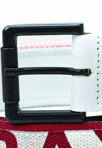 Boucle en métal noir de forme rectangulaire, fixée à une sangle blanche texturée, avec des lettres rouges et des détails de couture visibles.