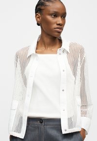 Νεαρή γυναίκα με πλεξούδες, φοράει λευκό πουκάμισο με τρυπήματα πάνω από λευκή μπλούζα και σκούρο γκρι παντελόνι σε απλό φόντο.