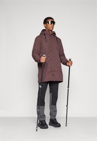 Veste d'extérieur violette avec une capuche, bâtons de trekking noirs et pantalon gris et noir. La veste est dotée de zips et de poignets réglables.
