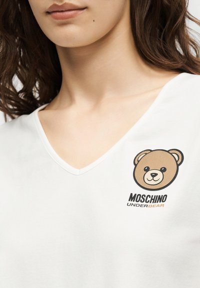Γυναίκα που φοράει λευκό πουκάμισο με V λαιμόκοψη, με μικρό καφέ πρόσωπο αρκούδας και το κείμενο «Moschino Underbear» στο αριστερό στήθος.