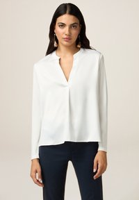 Blusa de seda blanca con escote en V y mangas largas, con un corte relajado y textura suave. Combinada con pantalones oscuros de color marino.