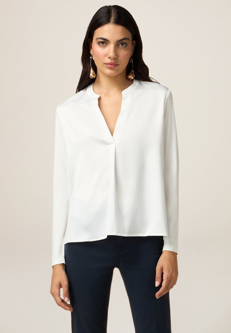 Blusa de seda blanca con escote en V y mangas largas, con un corte relajado y textura suave. Combinada con pantalones oscuros de color marino.