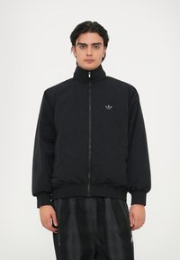 Crna podstavljena jakna s visokim ovratnikom i prednjim zatvaračem. Sadrži rebraste manžete i rub, s malim sivim logotipom na prsima.