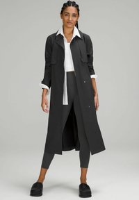 Długi czarny płaszcz trench z kołnierzem, noszony na białej koszuli i czarnych legginsach. Charakteryzuje się eleganckim designem i jednolitym kolorem, w połączeniu z czarnymi klapkami.