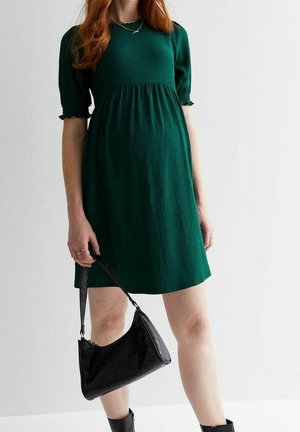 Kobieta w krótkiej sukience o ciemnozielonym kolorze, trzymająca czarną torebkę, stojąca na tle gładkiego białego tła.