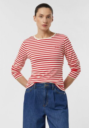 Frau trägt ein rot-weiß gestreiftes Langarmshirt und hoch taillierte blaue Jeans, steht mit den Händen am Rücken.