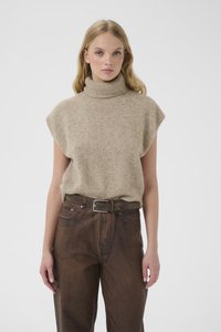 Junge Frau mit blonden Haaren, die einen beigen, ärmellosen Rollkragenpullover und braune, hoch taillierte Hosen mit einem Gürtel vor einem weißen Hintergrund trägt.