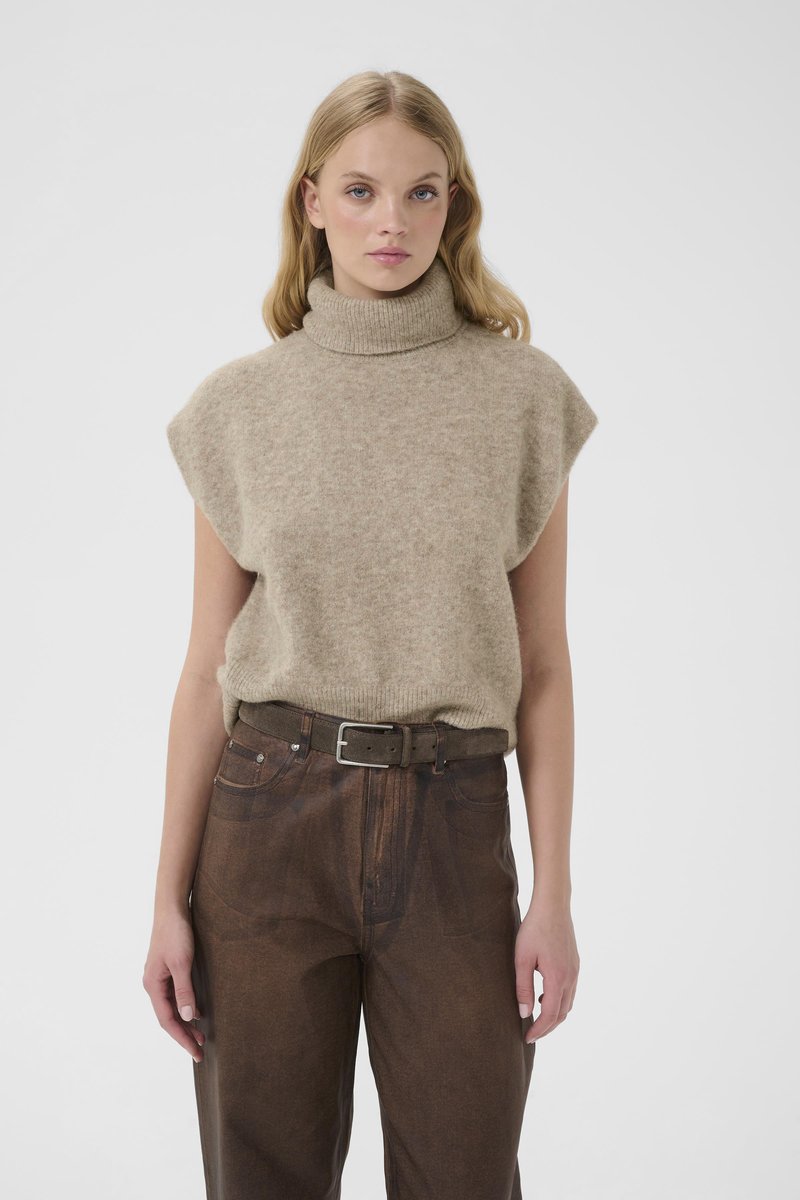 Junge Frau mit blonden Haaren, die einen beigen, ärmellosen Rollkragenpullover und braune, hoch taillierte Hosen mit einem Gürtel vor einem weißen Hintergrund trägt.