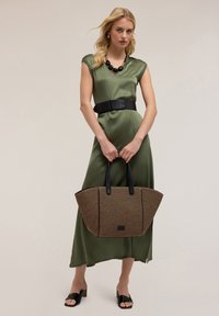 Abito in satin verde oliva con maniche a cappuccio, cintura larga nera e una grande borsa a tracolla intrecciata con manici e dettagli neri. Sandali neri con tacco basso.