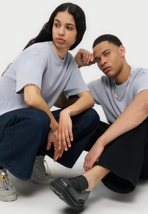 Δύο νεαροί ενήλικες με ανοιχτόγριζα πουκάμισα και σκούρα παντελόνια με φαρδιά πόδια, κάθονται κοντά στο πάτωμα μπροστά σε απλό φόντο.