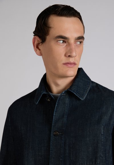 Mężczyzna o krótkich ciemnych włosach, ubrany w ciemną kurtkę dżinsową, patrzący w prawo na tle prostej, jasnoszarej ściany.