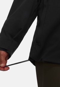 Svart vattentålig jacka i tyg med justerbar dragsko i nederkanten. Närbild av hand som håller i dragskon. Slät yta.