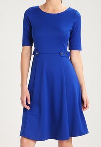 Robe A ligne bleu en tissu lisse, dotée de manches courtes, d'un col rond, et de deux accents de boutons à la taille.