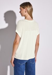Hvid kortærmet top med struktureret zigzag-mønster på ryggen, glat front og opkørte ærmer, parret med blå denimjeans.