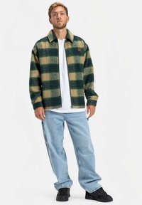 Πράσινο και μπεζ καρό μπουφάν από φλις με γιακά, κλείσιμο με φερμουάρ και καφέ λογότυπο, συνδυασμένο με ανοιχτό μπλε φαρδιά τζιν και μαύρα παπούτσια.